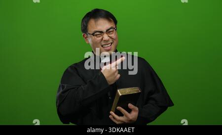 Asian man in clerical attire holding a book and gesturing against a solid green background, displaying a thoughtful expression. Stock Photo