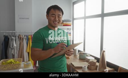 Asian man volunteering in a charity center, wearing a green shirt, taking notes on a clipboard in a room full of donations. Stock Photo