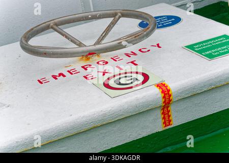 A metal hatch labeled emergency exit has a round turning handle and an unbroken security seal tape strip, showing the exit is closed and monitored on Stock Photo