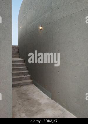 Architectural view of a narrow concrete stairway leading up between tall curved grey walls with a small light fixture, minimalist design. Stock Photo