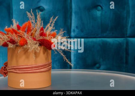 A flower arrangement with orange and beige tones sits in a pot on a table in front of a blue wall. Stock Photo