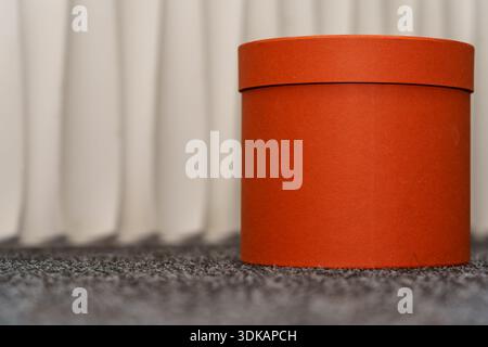 A round orange box is placed on flat gray carpet beside white curtains. Soft light fills the room creating a warm space. Stock Photo