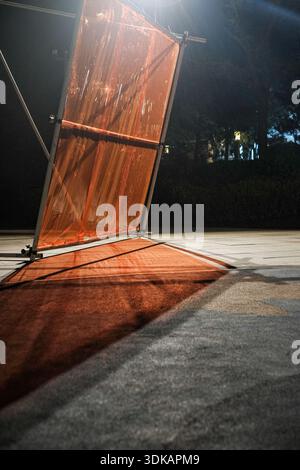Nighttime vertical shot of a temporary orange mesh barrier or safety screen illuminated by strong artificial light, casting reflections on the pavemen Stock Photo