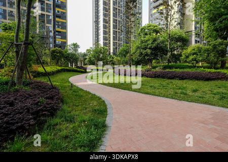 A curving red brick pathway winding through a green lawn and trees within a modern housing estate park. Stock Photo