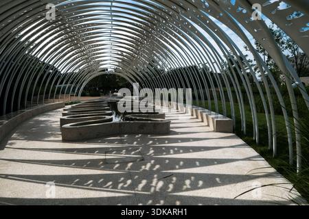 Horizontal shot of a long, curved white steel pergola structure in a park, showing the interplay of light and shadow on the paved ground and surroundi Stock Photo