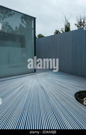 Vertical architectural shot of a building corner featuring a glass wall, a grey textured wall, and a floor with radiating blue and white striped lines Stock Photo