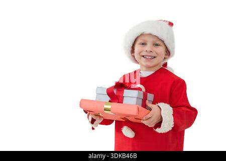 Schoolboy wearing Santa coat and hat, holding red and silver gifts in studio, copy space. Boy, festive, red, presents, wrapped, backdrop, white Stock Photo