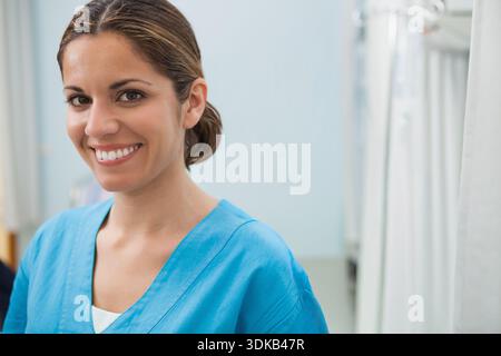 Female medical worker standing and smiling in clinic wearing blue scrubs near privacy curtain. Nurse, portrait, hospital, professional, cheerful, curt Stock Photo