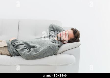 Man reclining on light-gray leather sofa at home, wearing knit sweater, head on armrest, copy space. Relaxed, minimalist, monochrome, lounge, serene, Stock Photo