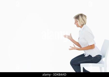 Senior woman is sitting on chair at right of backdrop, wearing white shirt, jeans, copy space. Elderly, speaker, palms, gesturing, presentation, studi Stock Photo