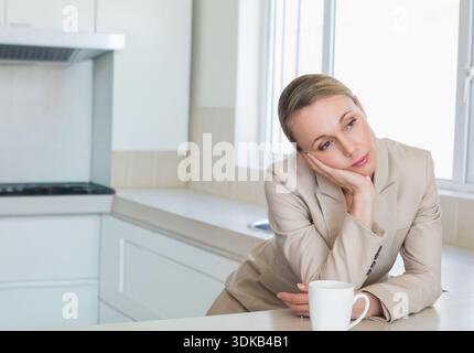 Mid adult woman wearing blazer leaning on counter in kitchen, resting head, ceramic mug, copy space. Contemplative, professional, sunlit, minimalist, Stock Photo