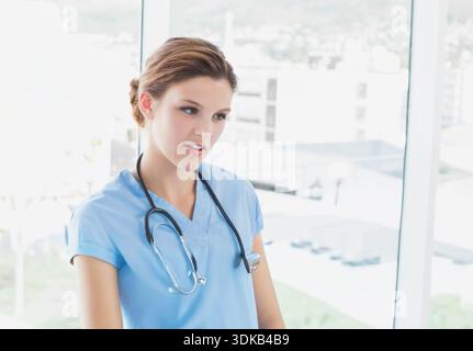 Female nurse in twenties standing in clinic near window wearing light blue scrubs stethoscope badge. Clinician, hospital, daylight, urban, greenery, c Stock Photo