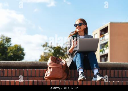 Modern woman with smartphone and laptop sitting on urban steps Stock Photo