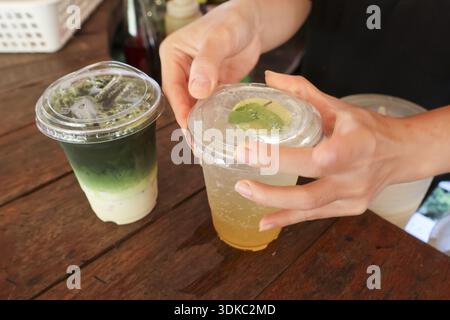 Person hand closing cold refreshing beverage cup on wood table. Another matcha green tea drink sits nearby, perfect moment of enjoyment at local cafe Stock Photo