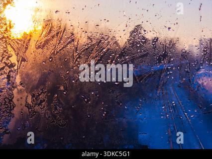 Window pane showing intricate ice patterns and water droplets reflecting a warm golden sunrise on one side and a cool blue twilight on the other, crea Stock Photo