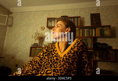 Young woman in a yellow leopard print sweater looking intently towards the side, thinking in a dimly lit room with bookshelves and floral wallpaper, c Stock Photo