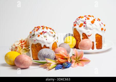 Two beautifully decorated Easter cakes, known as kulich, are presented with vibrant dyed eggs and delicate spring flowers on a clean white background Stock Photo