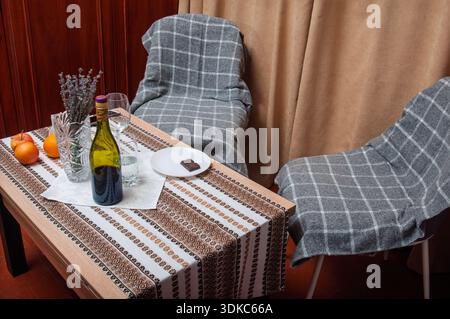 A bottle of wine, fruit, and chocolate are arranged on a table with a decorative runner, set for two with chairs draped in blankets Stock Photo