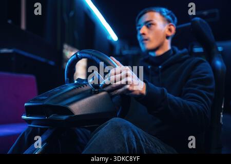 In the seat. Young guy is playing racing game by using driving simulator. Stock Photo