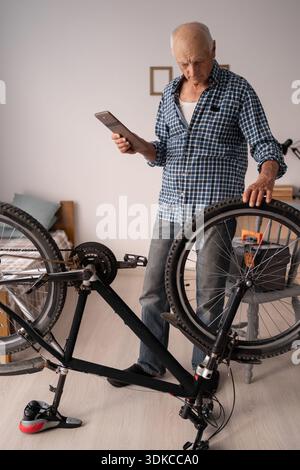 Elderly man inspecting and repairing his bike using digital tablet at home. Senior male cyclist maintain bicycle. copy space Stock Photo