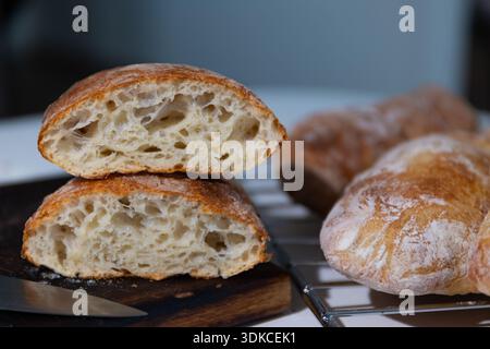 Two halves of rustic white bread rest on a dark wooden cutting board, revealing a light, open crumb. Other golden-crusted loaves cool on a metal rack nearby, freshly baked in a home kitchen. Stock Photo