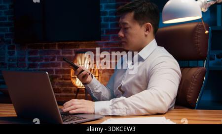Serious Asian 40s business man recording audio message talk mobile phone at night evening office Korean businessman record voice mail talking to Stock Photo