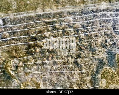 Frozen water surface featuring naturally formed ice lines and delicate crack patterns reveals the rocky bottom. Translucent ice creates unique texture. Stock Photo