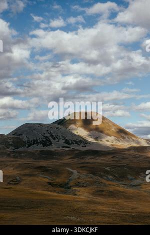 Sunlit volcanic peak rising from golden highlands under dramatic moving clouds in a vast, quiet mountain wilderness. Stock Photo