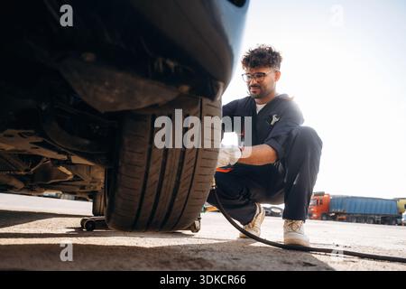 Sitting, side view, changing tire, using screwdriver. Mechanic is with the car outdoors. Stock Photo