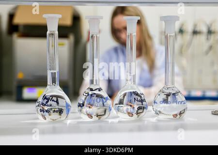 Volumetric laboratory flasks containing clear liquid on a workbench, with a female scientist working in the background Stock Photo