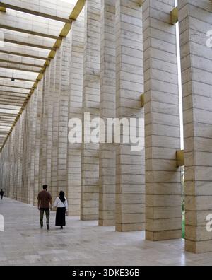 A couple walks hand-in-hand through a grand hall featuring a row of massive, light-colored stone pillars Stock Photo