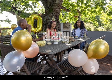 Diverse friends sitting at table on deck beside balloons wearing party hats and eating strawberries. Celebration, togetherness, outdoor, rustic, cheerful, socializing, festive || Model released Stock Photo