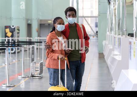 Portrait of millennial black couple in medical protective mask hold boarding pass and passports in airport. African American young tourists fly on vacation wait for plane departure in empty terminal || Model released Stock Photo