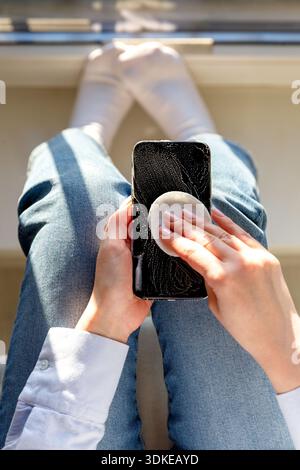 Woman cleaning mobile phone to eliminate germs, bacterias. Female disinfects a smartphone by applying sanitizer, alcohol disinfecting foam using a cotton pad, close up. Top view. || Model released Stock Photo