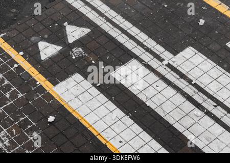 A close-up top view of a dark tiled sidewalk featuring bold white arrows and lines, accented by a yellow edge. Light snow rests on the surface, evokin Stock Photo
