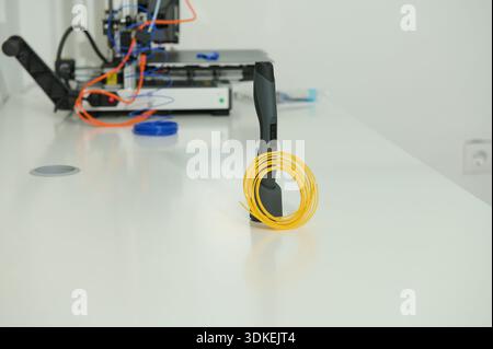A clean tech workspace features a yellow coiled cable wrapped around a handheld device on a white workbench, with circuitry gear and a 3D printer in t Stock Photo