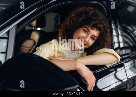 Looking through the opened window and smiling. Happy woman is sitting in the car. Stock Photo