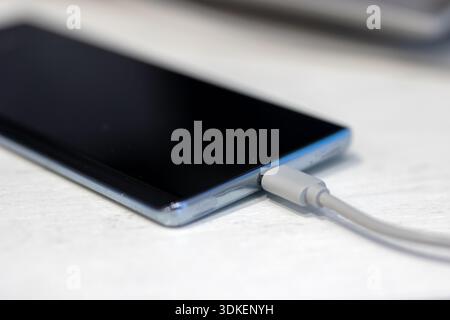 Close up smartphone charging on table, white cable plugged into port, glossy screen reflecting soft daylight, shallow Stock Photo
