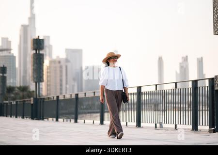 young woman walking in the city with tall buildings in background at sunset, waterfront evening walk Stock Photo