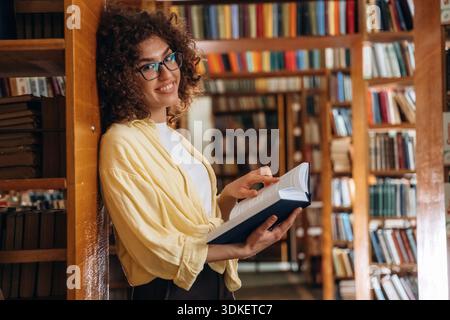 Standing and leaning on the shelf. Beautiful woman is in the library. Stock Photo