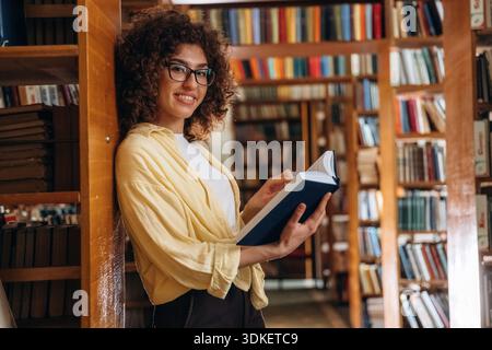 Standing and leaning on the shelf. Beautiful woman is in the library. Stock Photo