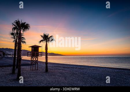 A tranquil sunset on a pebble beach features palm trees and a lifeguard tower overlooking a calm sea, Castell de Ferro, Spain Stock Photo