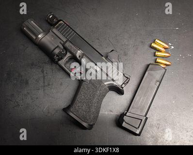Modern semi-automatic black handgun with tactical flashlight, optical sight, extended magazine and ammunition on dark textured background for defense Stock Photo