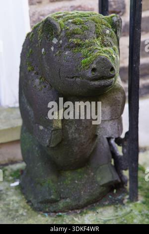 Moss-covered stone pig statue sitting and hugging its knees in a garden setting. London,England Stock Photo
