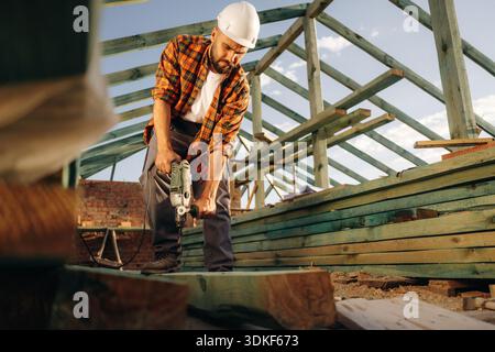 Drilling the holes in plank. Construction worker is on the roof of a house. Stock Photo