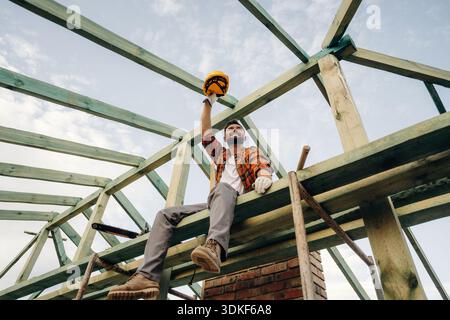 Friendly man is sitting and taking a break. Construction worker is on the roof of a house. Stock Photo