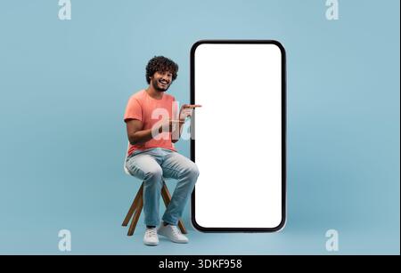 Positive indian guy in casual sitting by huge cell phone Stock Photo