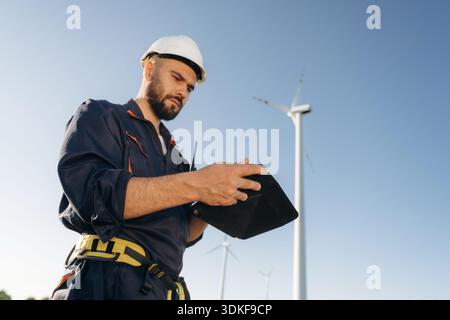 Reading information on digital tablet. Engineer in field checking on wind turbine production. Stock Photo