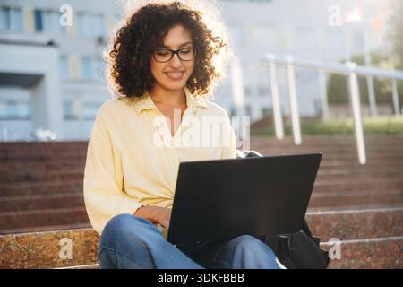 Using laptop. Beautiful woman student is near the university outdoors. Stock Photo