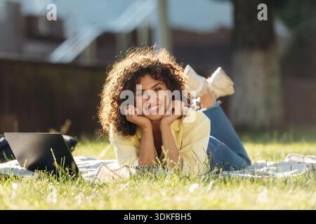 Smiling woman student is lying down on the green grass with laptop. Stock Photo